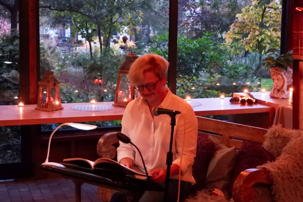 Właścicielka Centrum Ogrodniczego czyta Dzieło Izabeli Czartoryskiej „Myśli różne o sposobie zakładania ogrodów”. Za nią tło stanowią duże szyby, za którymi widać ogród w resztkach światła zachodzącego dnia