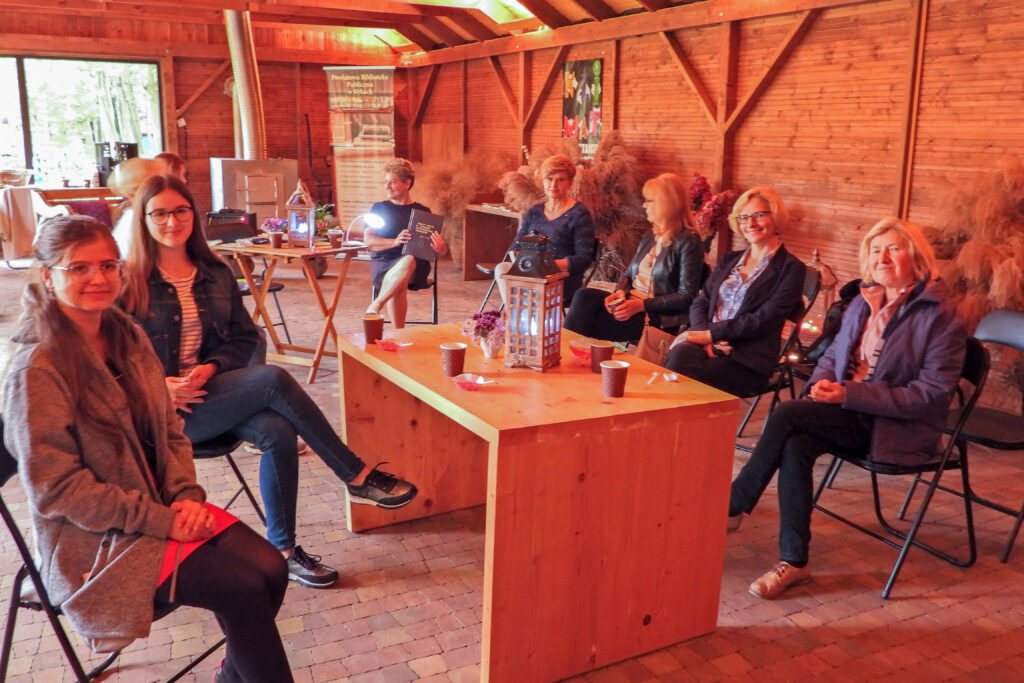 uśmiechnięta grupa ludzi siedzi wokół drewnianego stołu, prowadząca trzyma książkę, którą omawiają po czytaniu fragmentów, akcja dzieje się w Daglezji na Zielonej w Rykach z okazji Nocy Bibliotek w dniu 10.10.2020