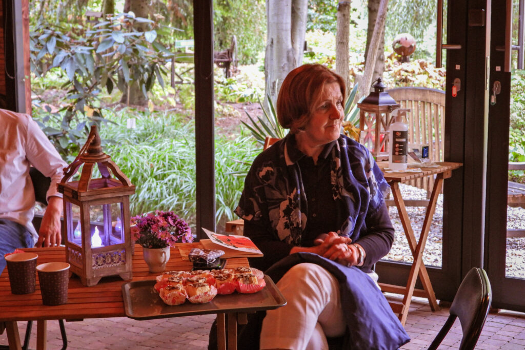 na pierwszym planie siedzi kobieta w rozpiętym ciemnym płaszczu, ubrana w jasne spodnie i fantazyjnie zarzuconą chustą na ramiona i płaszcz, założyła nogę na nogę i trzyma dłonie złożone na kolanach, twarz ma zamyśloną, słucha książki, jest zafascynowana. Obok niej z lewej strony stoi mały kwadratowy stoliczek, na którym stoi dużo ciasta powkładanego w kolorowe foremki papierowe mufinki, jest też ksiązka, w małym flakoniku piękne kwiaty jesienne, dwa kubki z kawą i duży lampion ze świecą. Za nią stoi drugi stolik, na którym widać płyn do dezynfekcji rąk, kolorowe karteczki przy wejściu i pojemnik z maseczkami też kolorowymi. W tle olbrzymie okno, z którym rozpościra się widok na ogród