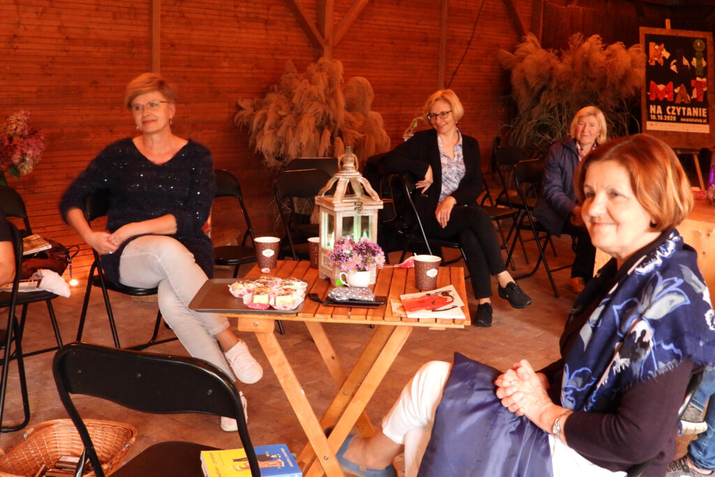 Na zdjęciu widać cztery kobiety, które patrzą w tę samą stronę, za nimi w dwu wielkich donicach naręcze długich traw ładnie wypełniających wnętrze, pomiędzy trawami stoi tablica z plakatem reklamujący nocne czytanie z treścią: klimat na czytanie 10.10.2020 r.