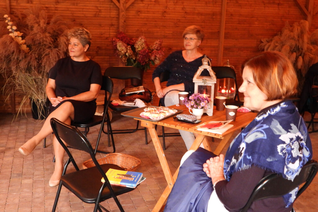 Na tle drewnianej ściany pośród ozdobnych traw siedzą trzy uśmiechnięte kobiety słuchając Dzieło Izabeli Czartoryskiej „Myśli różne o sposobie zakładania ogrodów”. Siedzą dookoła kwadratowego stolika, na którym stoją: ciasto, kawa, kwiaty, lampion ze świecą i telefon komórkowy, książka,