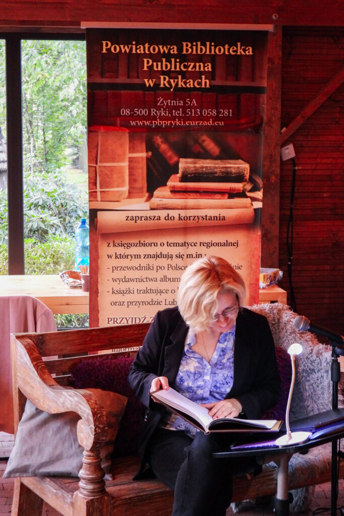 Kobieta w marynarce i niebieskiej bluzce w kwiaty pochyla się nad książka, jest rozbawiona, uśmiecha się i czyta. Siedzi na ławce pełnej miękkich poduszek, za nią rollup reklamujący biblioteke powiatową w rykach