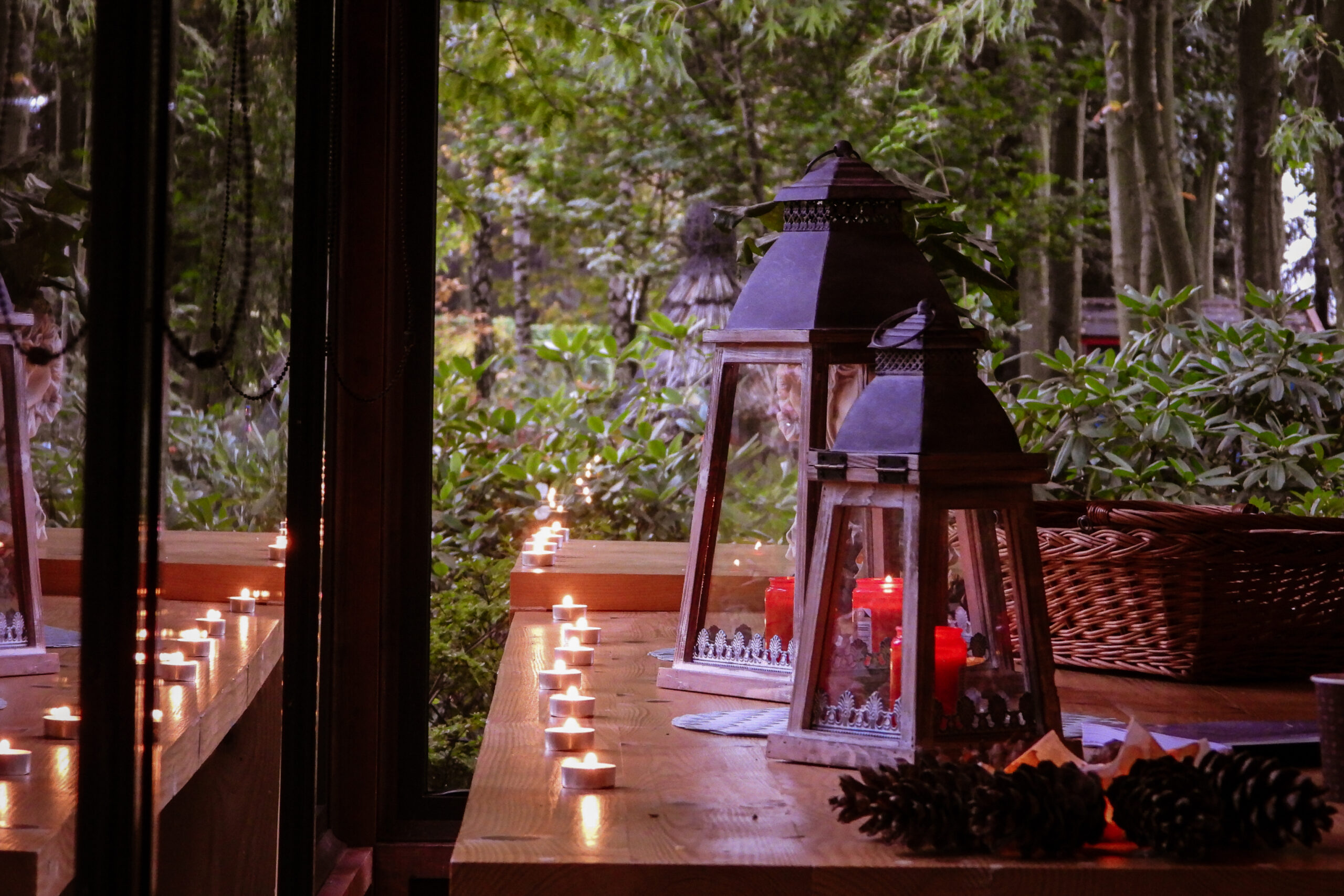 Zbliżenie na równo ułożony rząd świeczek na lakierowanym stole drewnianym, który pięknie odbija światło, po prawej dwie lapy z czerwonymi dużymi świecami olejowymi palącymi się spokojnym płomieniem, po lewej i z tyłu zamiast ścian dwie potężne szyby, za którymi ogród zaczyna tonąć w mroku. Wszystko pięknie wygląda dając prawdziwy klimat na nocne czytanie