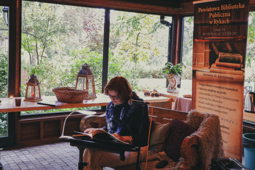 kobieta czyta w skupieniu siedząc na ławce pełnej poduszek. Za nią reklama biblioteki i w tle stoły w literę L a za nimi potężne okno z widokiem na ogród