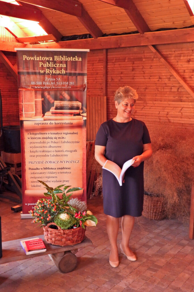 prowadząca to dyrektor Powiatowej Biblioteki Publicznej w Rykach, stoi na środku sali, z jej prawej strony rollup reklamujący bibliotekę, u stóp stoi zgrabna mała drewniana ławeczka na której w koszu wiklinowym ułożono dużo kolorowych kwiatów, obok ksiązki na ławeczce, wszystko na tle drewnianej ściany, którą przełamuje olbrzymia donica wypełniona miskantem, rodzajem bardzo zdobnej wysokiej trawy