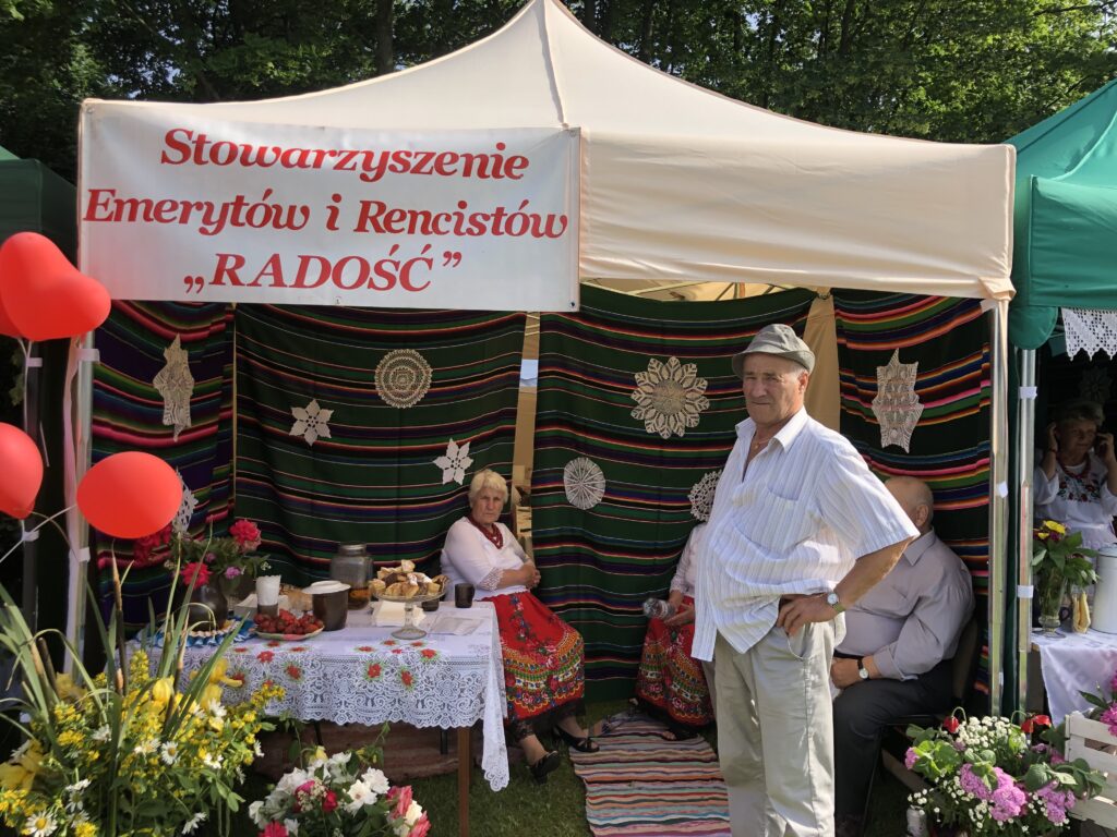 Stoi starszy pan na tle namiotu z napisem Stowarzyszenie Emerytów i rencistów Radość