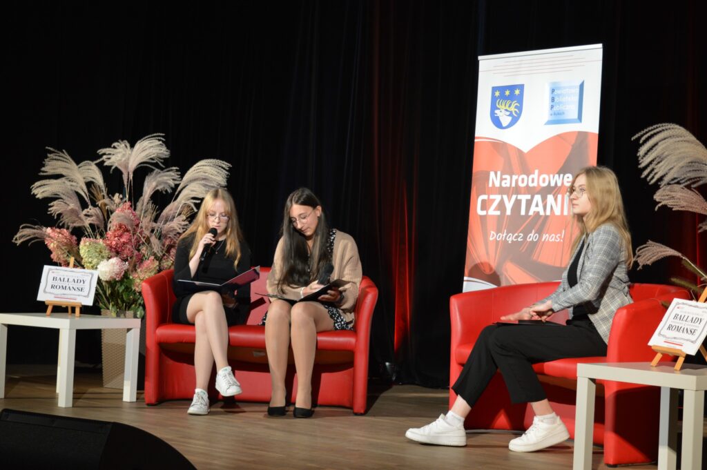 3 uczennice czytają na scenie. Ciemne tło, w środku banner mówiący o Narodowym czytaniu. Siedzą na czerwonych sofach