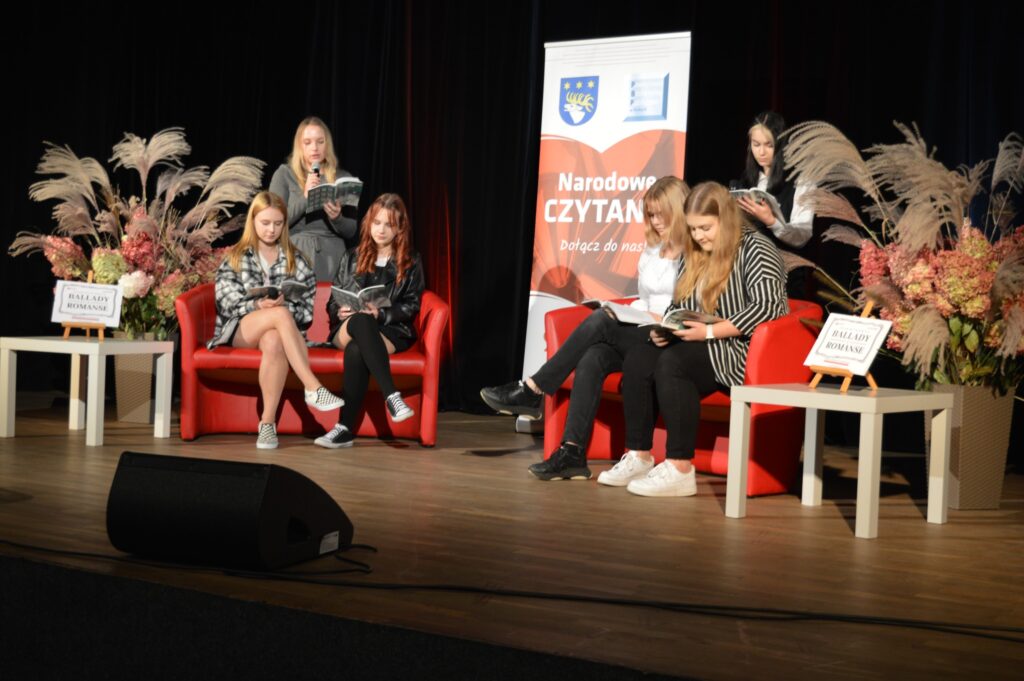 6 uczennic czyta na scenie. Ciemne tło, w środku banner mówiący o Narodowym czytaniu. Siedzą na czerwonych sofach