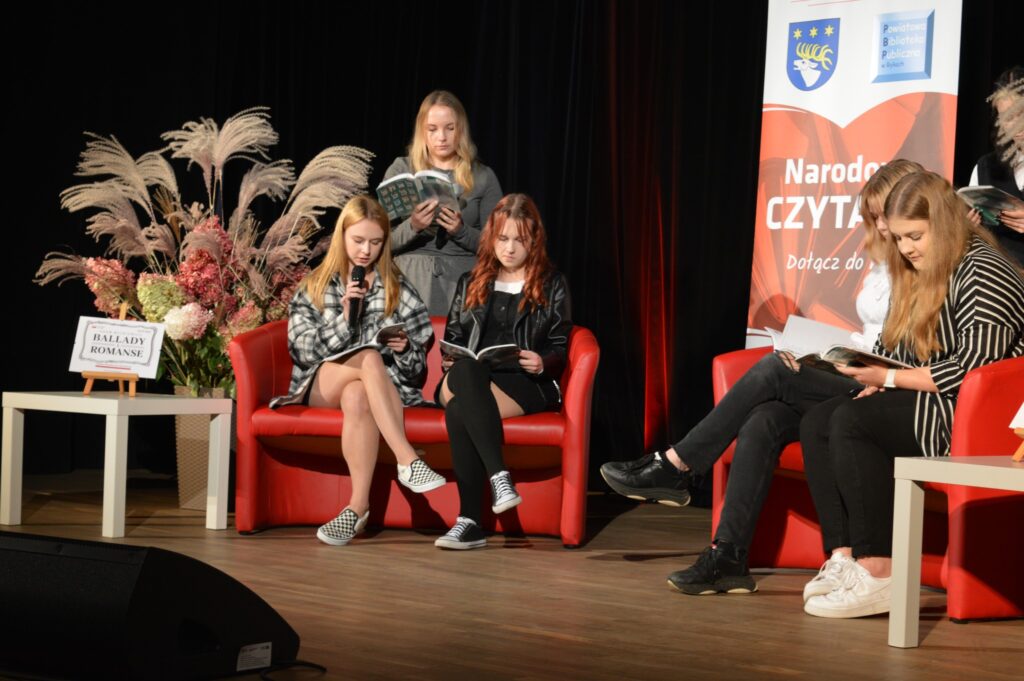 3 uczennice czytają na scenie. Ciemne tło, w środku banner mówiący o Narodowym czytaniu. Siedzą na czerwonych sofach