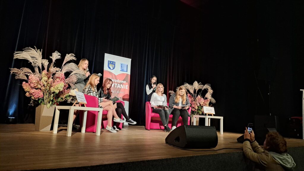 6 uczennic czyta na scenie. Ciemne tło, w środku banner mówiący o Narodowym czytaniu. Siedzą na czerwonych sofach, 2 stoją. Z przodu dyrektor biblioteki nagrywa ich komórką