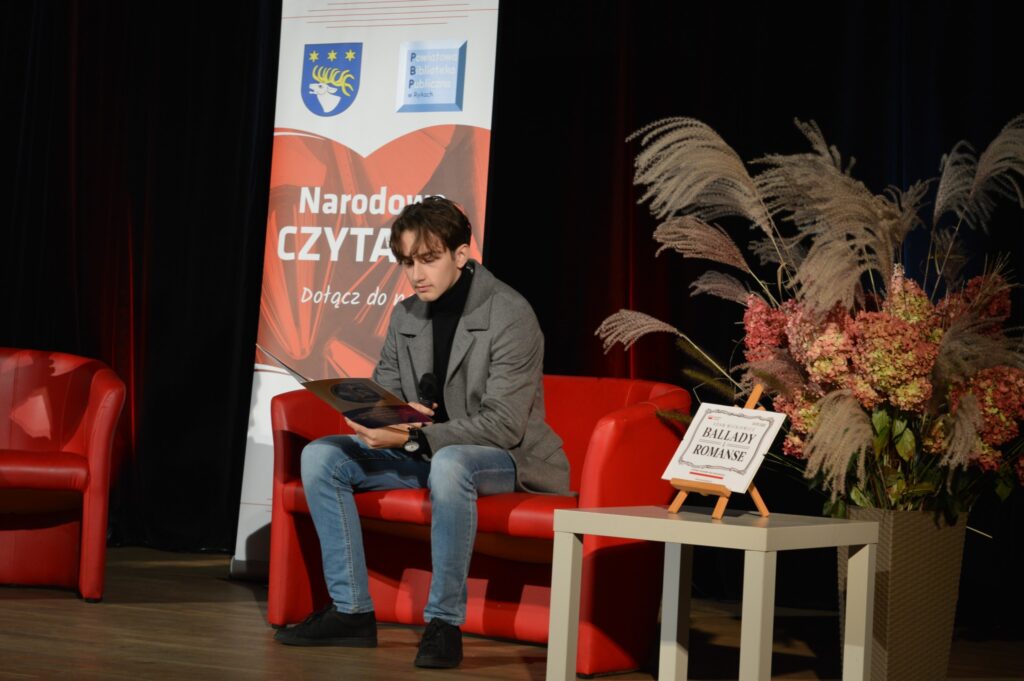 z tyłu baner reklamujący Narodowe czytanie. Wszystko na czarnym tle w kinie na scenie. Na pierwszym planie siedzi na czerwonej sofie chłopak i czyta,. Obok niego stolik z napisem Ballady i romanse, za nim potężny bukiet kwiatów ozdobiony piuropuszami traw