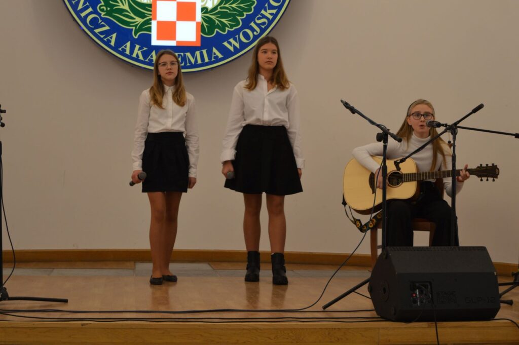 Przegląd Piosenki Patriotycznej i Harcersskij Powiatowej Biblioteki Publicznej w Rykach, scena, dwie dziewczyny śpiewają jedna gra na gitarze