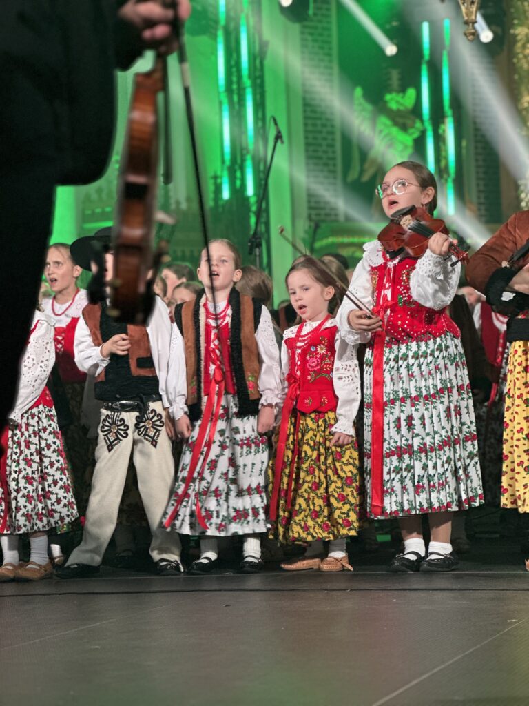 skrzypek w świetle reflektorów dyryguje zespołowi Mała Armia Janosika, dzieci śpiewają ubrane w góralskie stroje