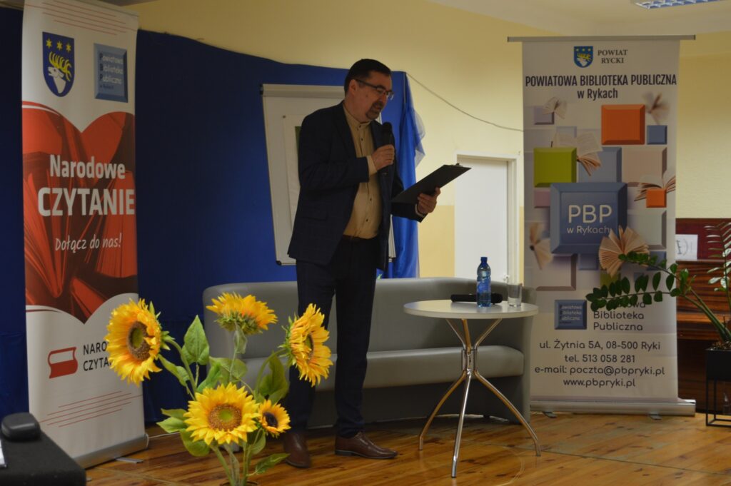 Czyta mężczyzna, czarny garnitur, stoi na scenie z boku żółte słoneczniki, tło narodowe czytanie, scena