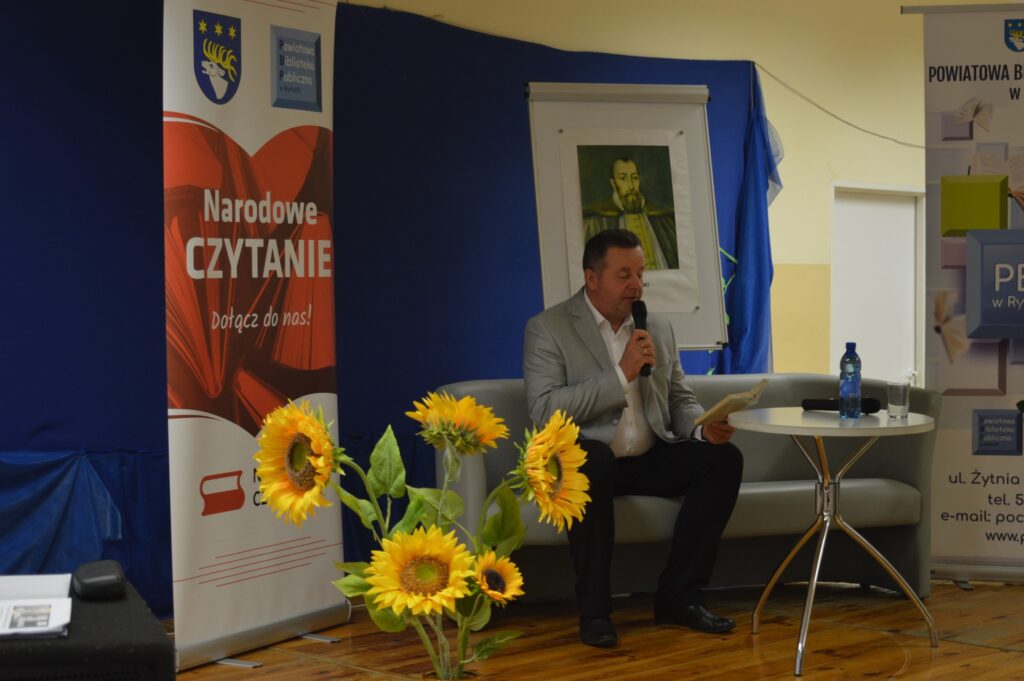 Czyta dyrektor ZSZ nr 1, siedzi, z boku żółte słoneczniki, tło narodowe czytanie, scena