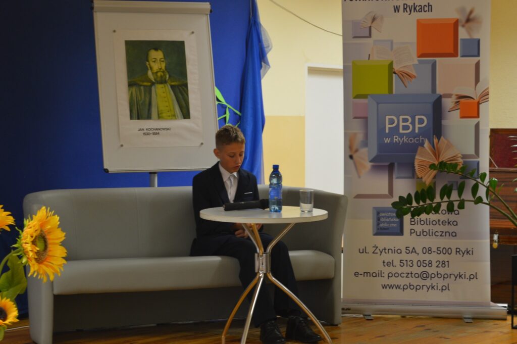 Czyta chłopiec, czarny garnitur, siedzi, na scenie z boku żółte słoneczniki, tło narodowe czytanie, scena