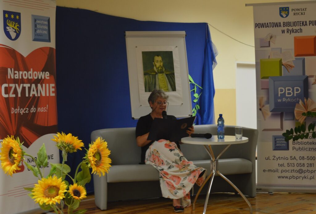 Dyrektor PBP Jarosz Dagmara  siedzi, czyta, z boku żółte słoneczniki, tło narodowe czytanie, scena