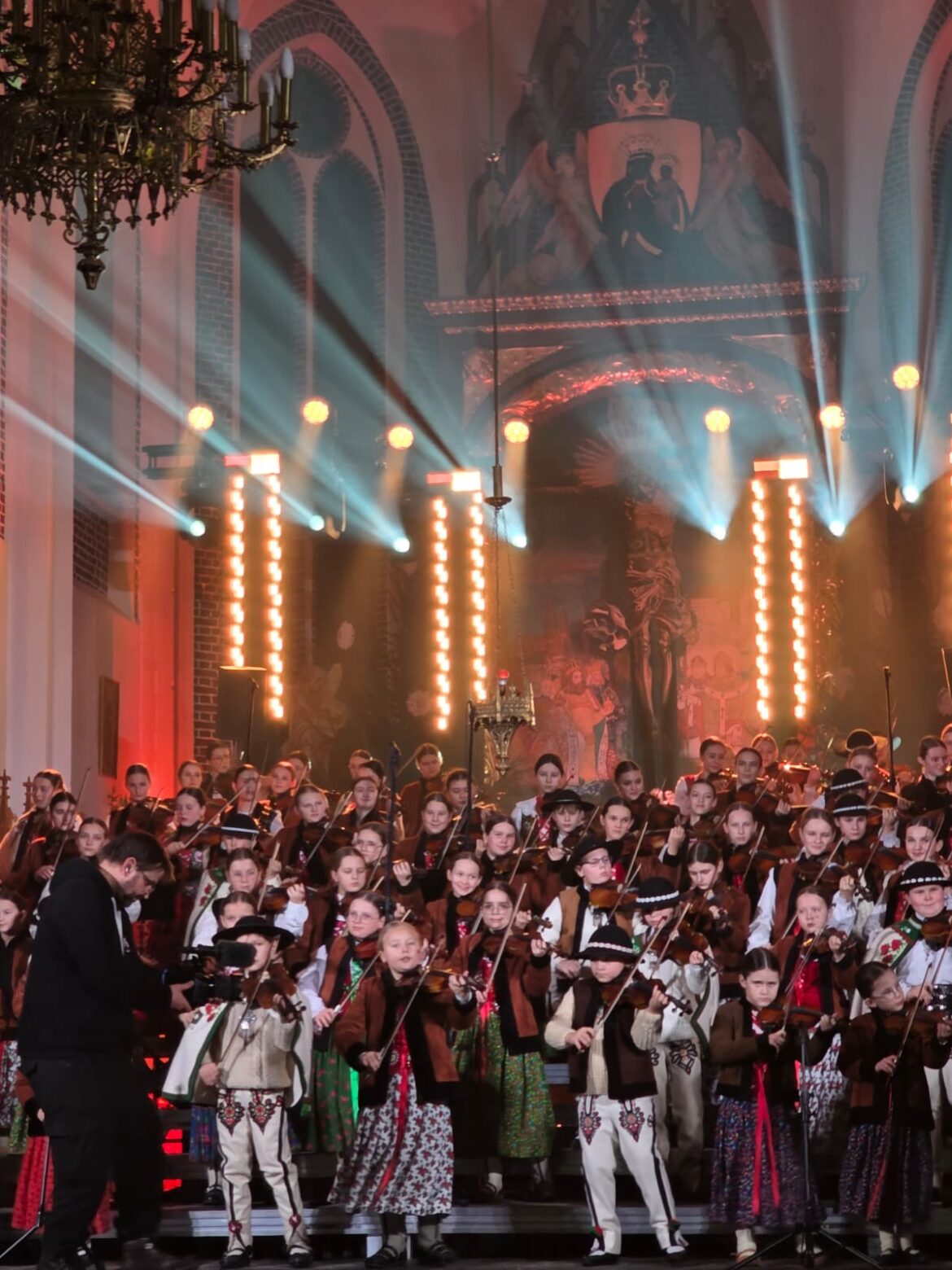 Koncert małej armii Janosika sceneria w Kościele, gra świateł, grupa dzieci w strojach góralskich a z lewej strony kamerzysta filmujący grających na skrzypcach